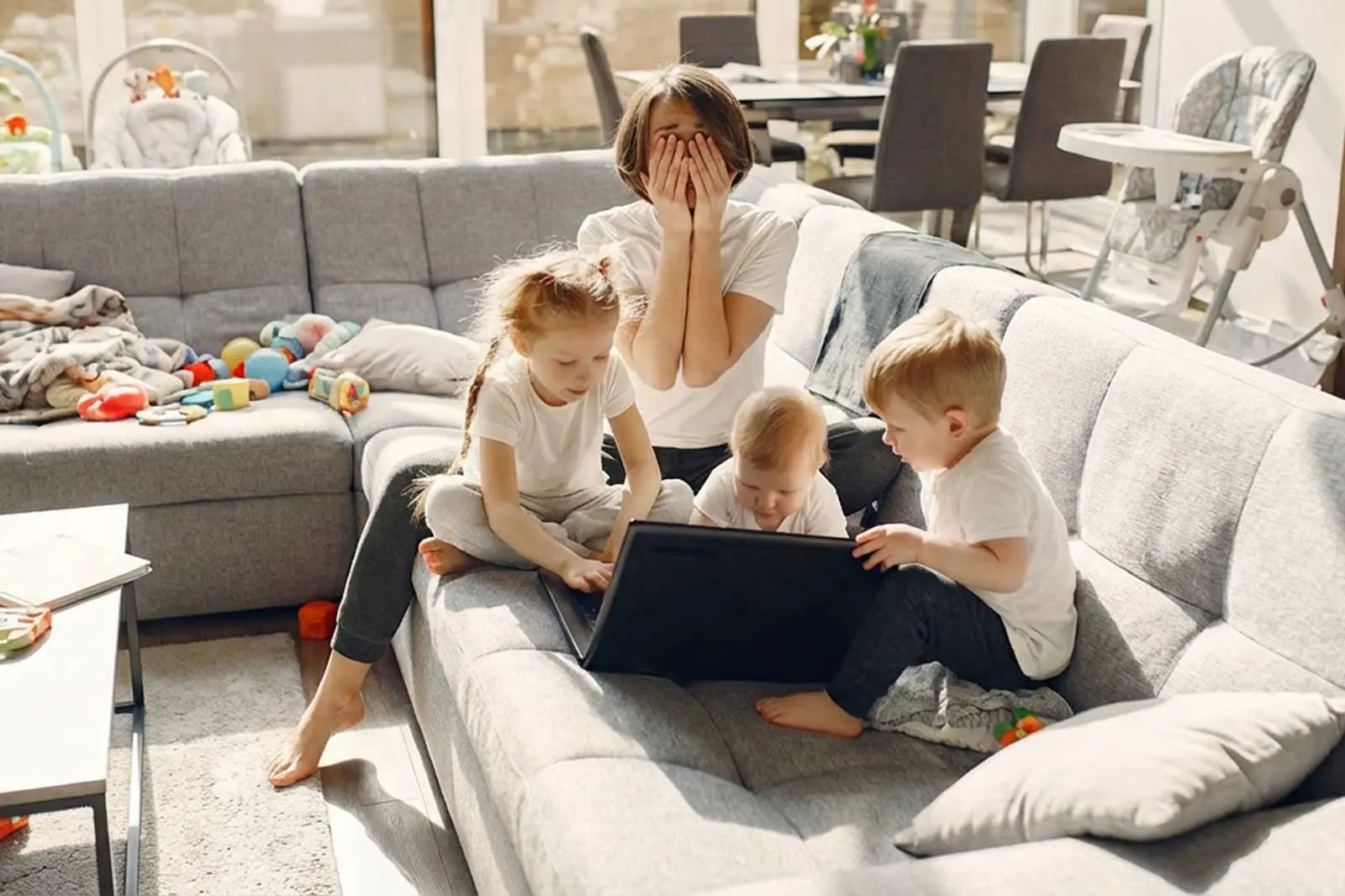 Madre abrumada cubriéndose el rostro sentada en un sofá rodeada de tres hijos pequeños y juguetes, representando el agotamiento parental.